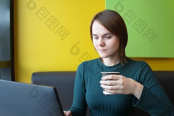 一个脸上带着沉思的平静表情的女孩坐在咖啡馆桌子旁的笔记本电脑旁，棕色沙发靠着黄色的墙壁。他看着电脑显示器，手里拿着一个纸杯，里面放着咖啡。