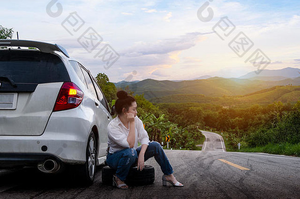 一位年轻美丽的女士坐在汽车旁呼救，在山林和天空背景下的公共道路上