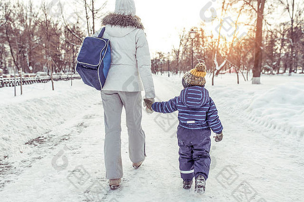 冬天，妈妈带着儿子在雪堆中的公园里散步后回到了家。牵着小男孩的手。