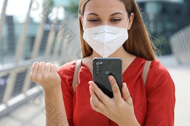 一位手持KN95防护面罩、手持拳头的年轻女子在手机上读着一条短信，表达了对这条好消息的喜悦。情感特写