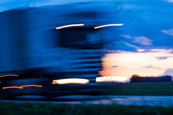 日落后，高速行驶的运动模糊了公路上亮着灯光的迎面驶来的卡车。照亮聚光灯下的汽车。黄昏时蓝橙色明亮的天空。