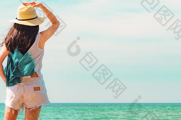 穿着休闲时尚的快乐年轻女子，戴着草帽站在海边。在翡翠色的热带天堂海滩放松身心，享受假期