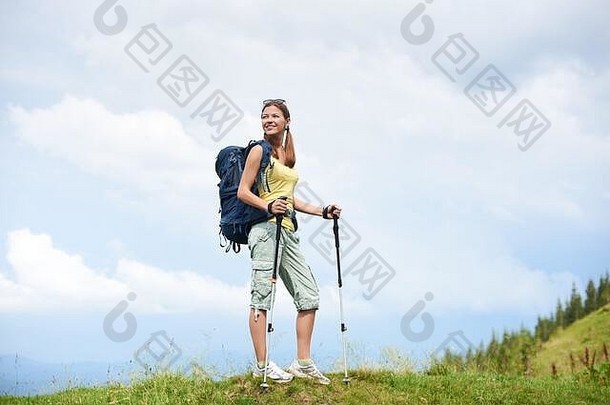 美丽快乐的女子徒步旅行者徒步走山路，走在青草丛生的小山上，背着背包，使用登山杖，享受夏日多云的天气。户外活动、旅游理念