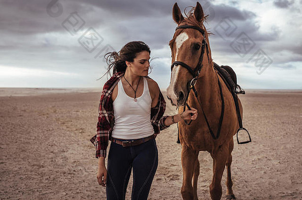 一个女人牵着一匹马在海滩上行走的肖像。晚上，女骑手和她的种马<strong>一起</strong>散步。