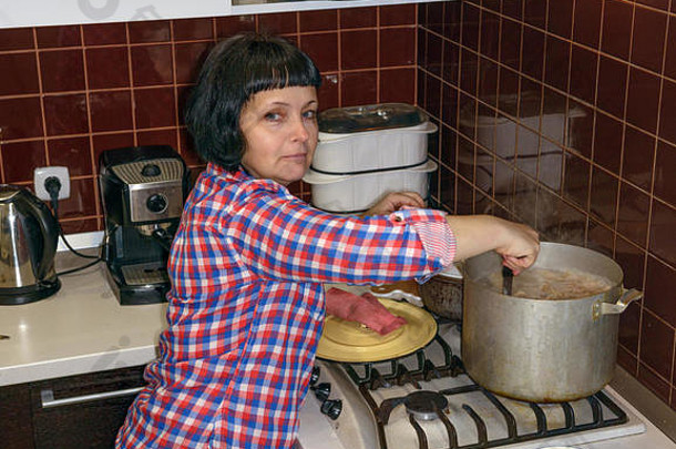 一位成熟的黑发女人正在家厨房里用大铝锅为她的看门狗煮粥。