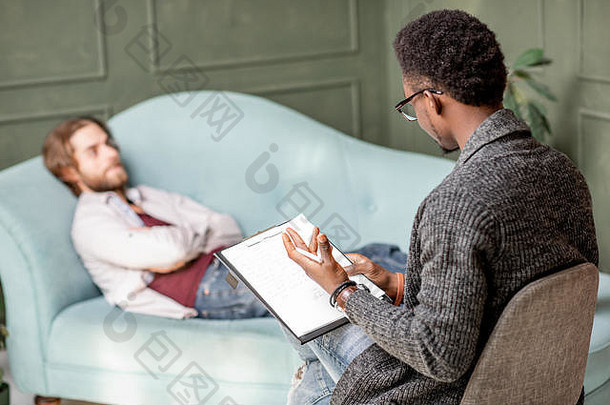 在绿色办公室里，一位年轻的白人男子正在拜访一位躺在舒适沙发上的非洲心理学家