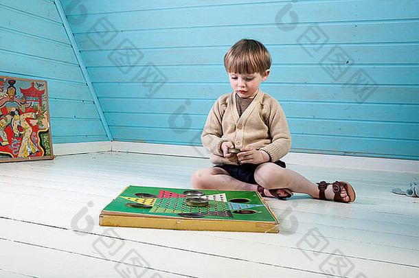 一个穿着漂亮衣服和凉鞋的小男孩在一间蓝白相间的阁楼里独自玩着老式的棋盘游戏。