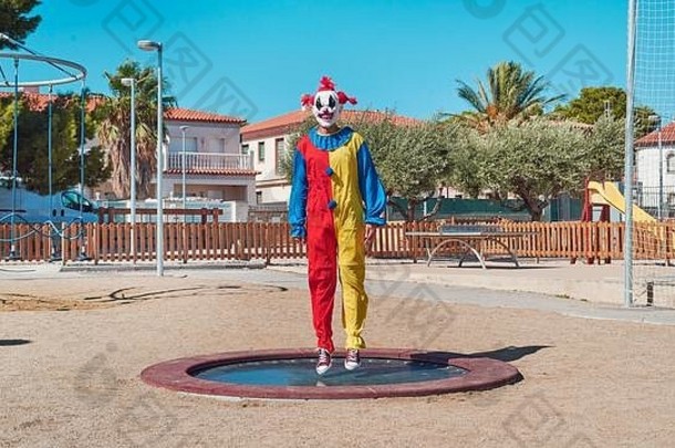 可怕的小丑穿色彩斑斓的黄色的红色的蓝色的服装跳跃蹦床户外公共操场上