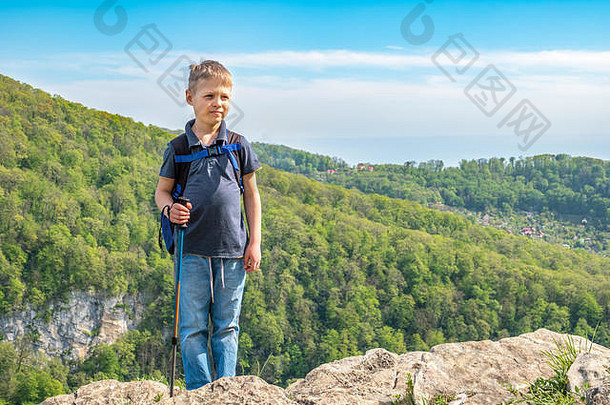 一个带着登山杖和背包的男孩站在绿色森林中的山顶上。山里春夏晴朗