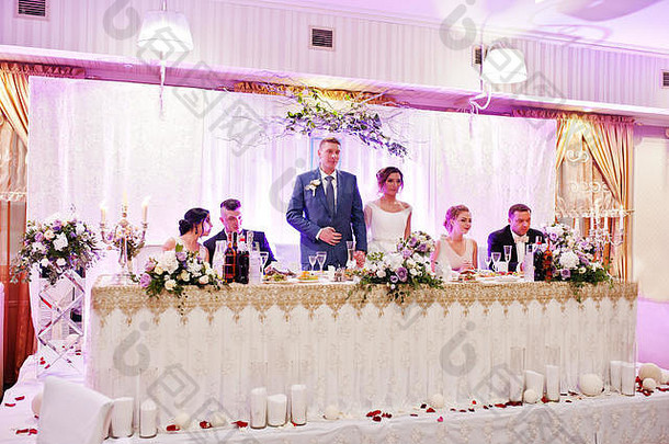 美丽的新婚夫妇站在桌旁，伴娘和伴郎坐在他们旁边。