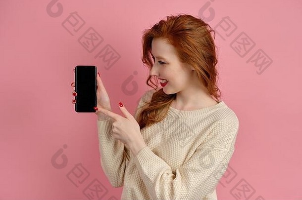 一个红头发的漂亮红发女孩站在粉红的背景上，戴着电话，微笑着指向黑暗的屏幕。
