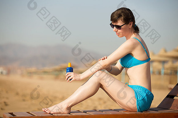 阳光明媚的海滩上，一位身穿蓝色泳衣、面带微笑的年轻女子，手里拿着一瓶防晒霜，在涂单宁之前将防晒霜涂抹在手臂上