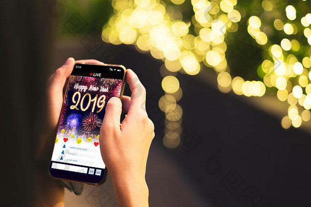 手持手机的女士观看2019年新年快乐的直播倒计时，屏幕上的焰火覆盖城市景观，背景为浅波基，Digi