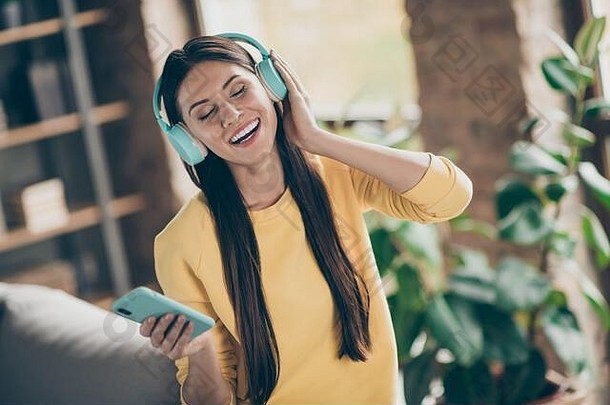 积极开朗女孩的肖像在智能手机上听音乐选择旋律欣赏歌曲在房间公寓穿黄色毛衣
