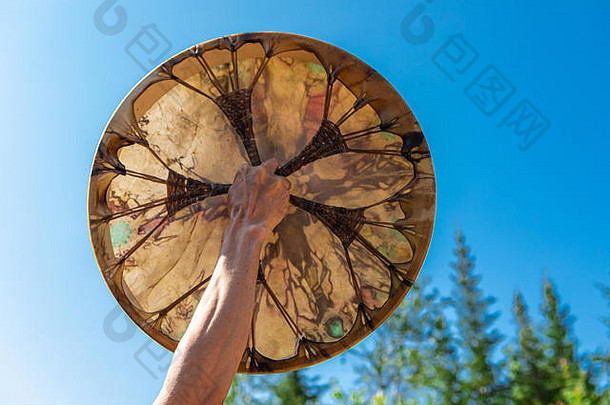 一名男子在晴朗的天空中手持他的神圣鼓，近距离地手持美国土著人的鼓，孤立地在蓝天和树木的背景下