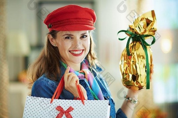 在阳光明媚的春日，一位身着牛仔裤衬衫、头戴红帽子、面带微笑、优雅的40岁老妇人在现代住宅中的画像，用金箔纸包裹着大e