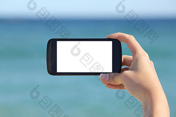 以大海为背景，在海滩上，一只女士的手在显示一个空白的智能手机水平屏幕
