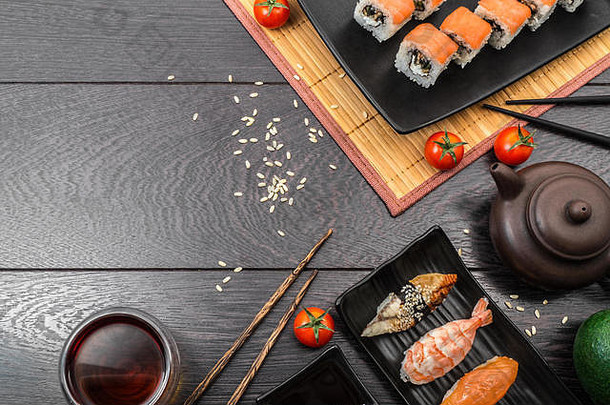 寿司<strong>套装</strong>生鱼片、尼吉里、马古罗和寿司卷放在<strong>盘</strong>子里，西红柿、茶壶和茶碗放在深色木质背景上。文本的空间。