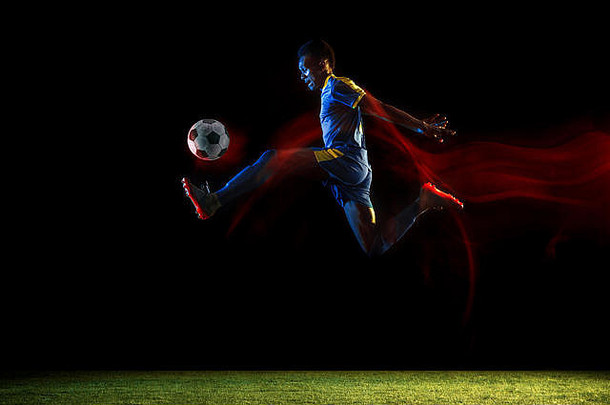 穿着运动服和靴子的年轻非洲裔美国男子足球或足球运动员，在黑暗背景的混合灯光下踢球。健康生活方式、职业运动、爱好的概念。