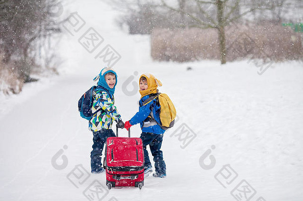两个小孩，背着背包和一个大红手提箱的男孩兄弟，去度假，在一个下雪的冬天在公园里散步