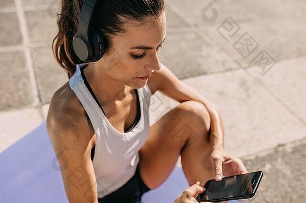 女人听音乐耳机聪明的电话锻炼女运动服装坐着在户外健身应用程序暴徒