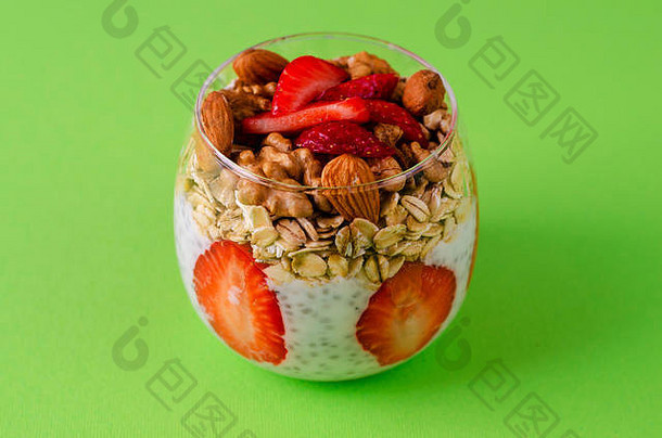 酸奶加布丁，配新鲜草莓、隔夜燕麦和坚果，置于绿色背景的玻璃杯中。前视图。均衡健康早餐理念。