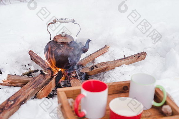浪漫的冬季野餐。两个杯子和一个碗放在雪地里的木制托盘上。背景上明火上方的铜壶模糊。<strong>烧水壶</strong>