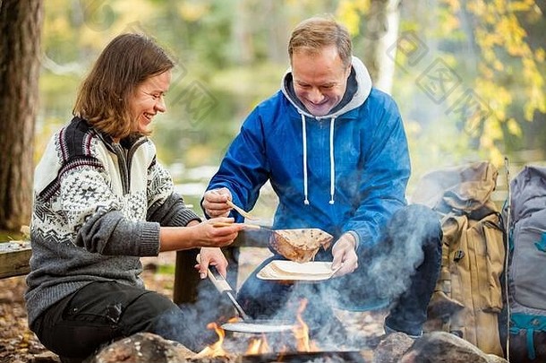 在湖边的森林里，一对男女在篝火上烤煎饼，生火，烧烤。探索芬兰的幸福夫妇。斯堪的纳维亚风景。