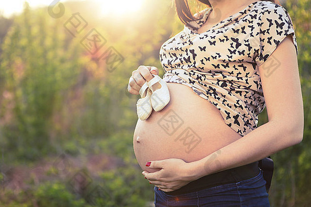 日出背景下腹部穿着婴儿小鞋子的无法辨认的孕妇的特写镜头