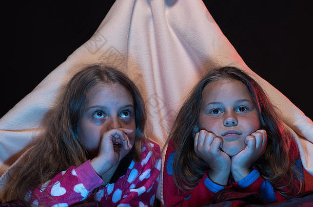 儿童和娱乐时间概念。有着复<strong>杂面</strong>孔的女孩。女朋友在毛毯帐篷里看电视。孩子们穿着黑色背景的红色睡衣躺在床上。儿童睡衣派对。
