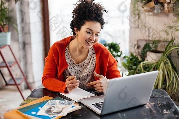 年轻的快乐的女人黑暗卷曲的头发坐着表格幸福的工作移动PC现代车间大窗户背景