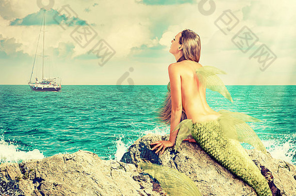 美丽的美人鱼带着鱼尾坐在岩石上，看着地平线上的一艘船