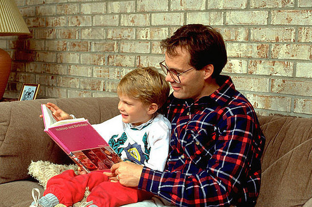 父亲30岁，儿子3岁，坐在客厅沙发上看书。美国明尼苏达州伯恩斯维尔