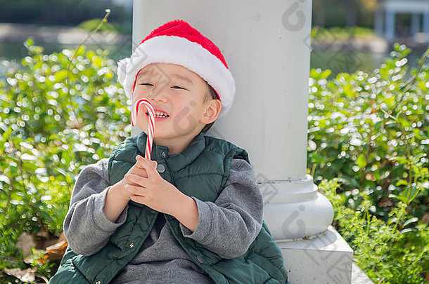 可爱的混血男孩坐在那里，戴着圣诞老人的帽子，享受着一根糖果手杖。