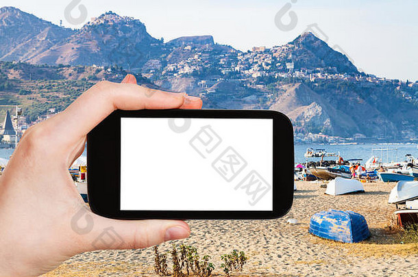 旅游概念-游客在夏天晚上用智能手机拍摄意大利西西里岛Giardini Naxos镇的城市沙滩和旧港口