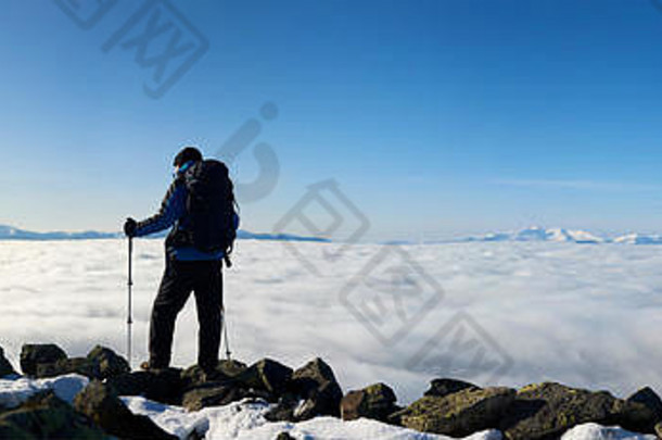 带着背包和<strong>登山杖</strong>的徒步旅行者站在山顶上，背景是黎明时的蓝天、云雾弥漫的山谷和远处的雪山。