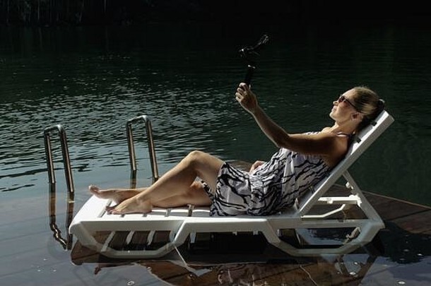 女子在手持胶片上拍摄智能手机万向节稳定装置。躺在码头上晒太阳的女孩自拍。博主播放视频博客。