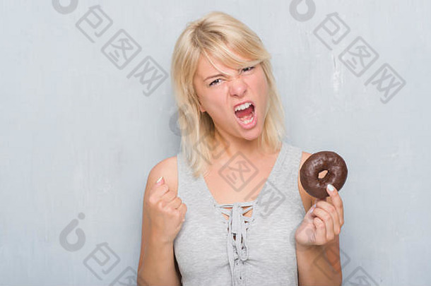 一位白人成年妇女在灰色的垃圾墙上吃着巧克力甜甜圈，恼怒而沮丧地大喊大叫，疯狂地举起手大喊大叫，ang