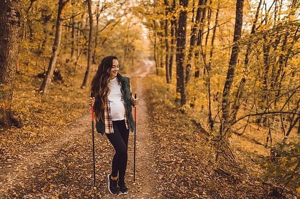 年轻快乐的孕妇带着登山杖在秋天的森林里徒步旅行。北欧步行和健身。怀孕活动和健康生活方式概念。