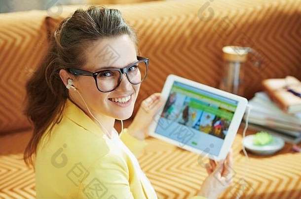 阳光明媚的日子里，穿着黄色夹克的快乐优雅的女人在现代的房子里通过平板电脑在线学习外语。