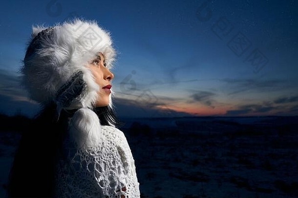 穿着时尚保暖冬季帽子和针织衬衫的美丽女孩的后视图。英俊的模特在冬日田野和夜空的背景下摆姿势。夜空中明亮的粉红色灯光。