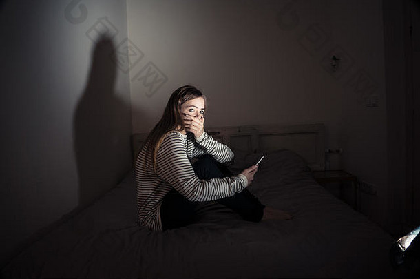 悲伤绝望的年轻女孩在智能手机上遭受在线欺凌和骚扰，晚上坐在床上感到孤独和绝望