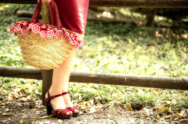 一位在公园里等候的妇女的修长而匀称的双腿，她穿着红色的管状连衣裙，<strong>高跟凉鞋</strong>，手里拿着一个乡村风格的装满鲜花的袋子