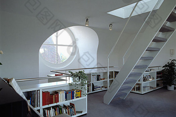 在现代白色阁楼转换客厅中，低书架上方的圆形窗户配有钢琴和楼梯