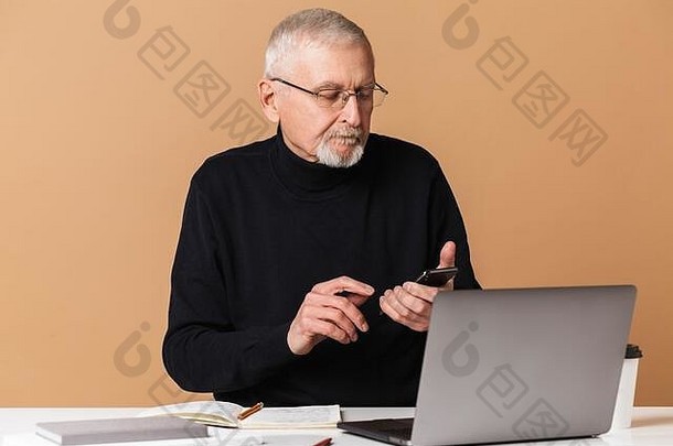 一位头发灰白、胡子灰白、戴着眼镜、穿着毛衣的帅哥坐在桌旁，手里拿着笔记本电脑、书和咖啡