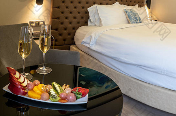 2个香槟酒杯，桌面上有一个水果碗，背景为床
