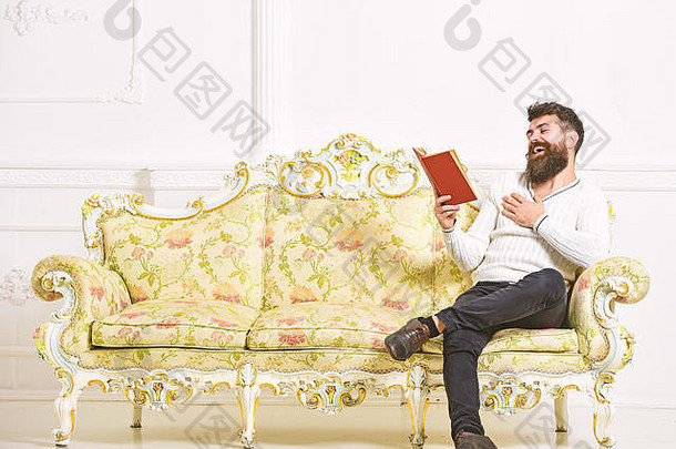 一个喜欢读旧书的家伙。留着胡子的男人坐在巴洛克风格的沙发上，拿着书，背景是白色的<strong>墙</strong>壁。男子气概的笑脸上读着书。幽默文学观。