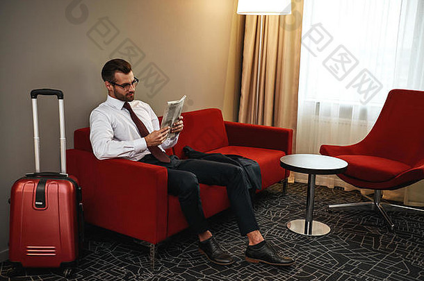 阅读<strong>最新新闻</strong>。戴眼镜的商人拿着手提箱和报纸坐在酒店大厅的沙发上