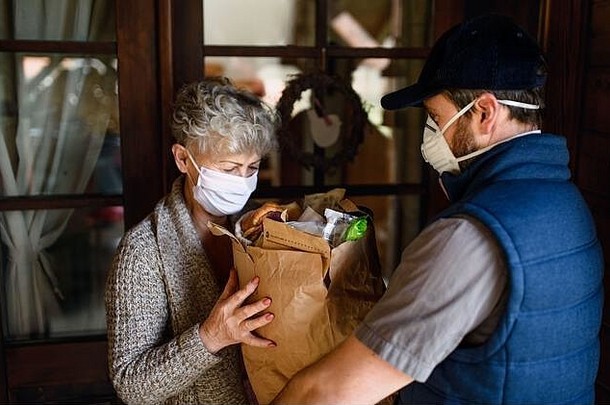 快递员带着口罩、电晕病毒和隔离概念为老年妇女运送购物物品。
