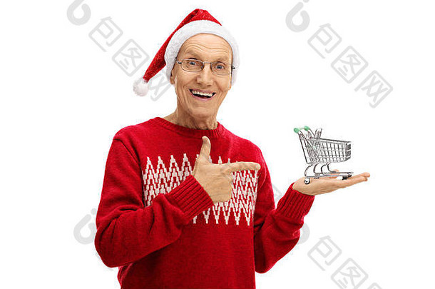 老人戴着圣诞帽，手里拿着一辆空的购物车，孤立地指着白色背景